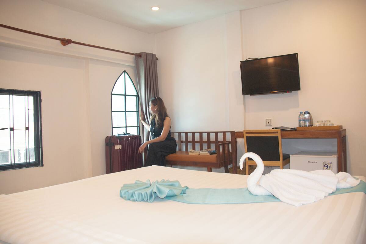 A hotel room with a bed, a TV mounted on the wall, a desk with a chair, and a window with curtains.