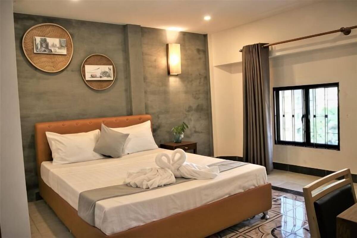 A bedroom with a large bed, a bedside table with a plant, a wall-mounted light, and a window with curtains.
