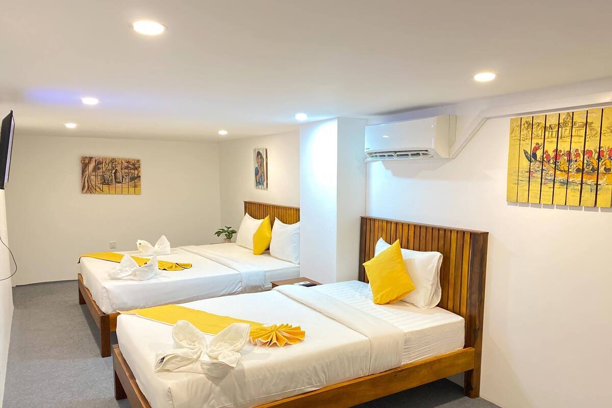 A hotel room with three single beds, each with white linens and yellow accents. There is a painting on the wall and an air conditioning unit above the beds.