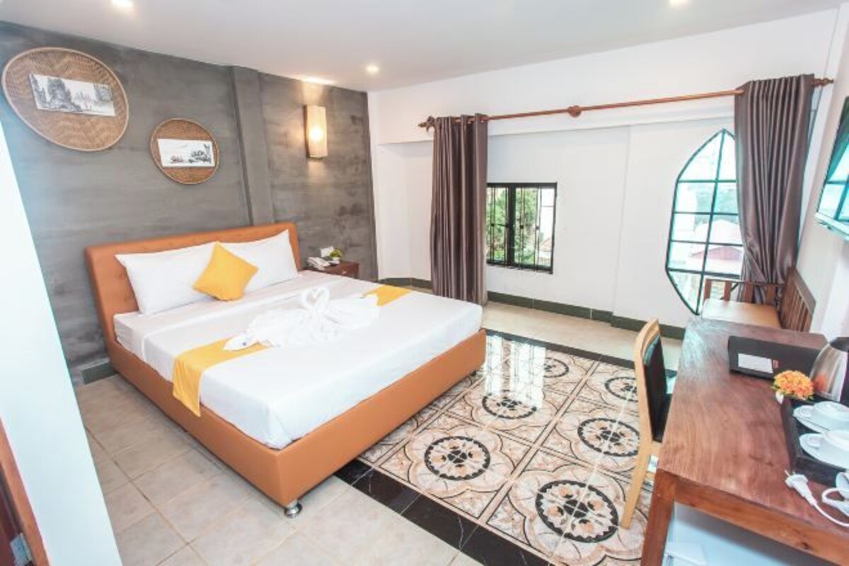 A hotel room with a bed, a desk, a chair, a window with curtains, and a wall-mounted light.