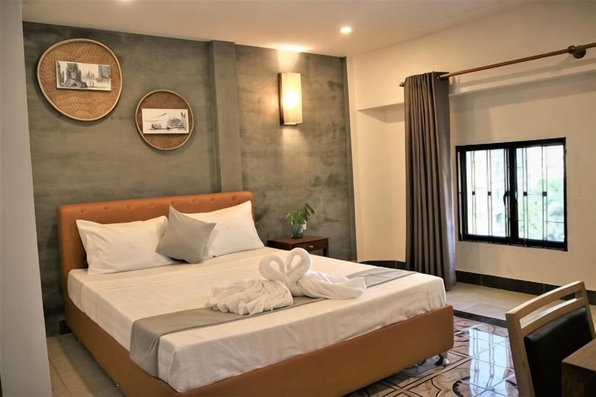 A bedroom with a large bed, a bedside table with a plant, a wall-mounted light, and a window with curtains.