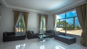 A modern living room with a sofa, armchair, and coffee table, large windows with curtains, and a view of greenery outside. - Saichon Grand View Resort (Ban Ta Khun)