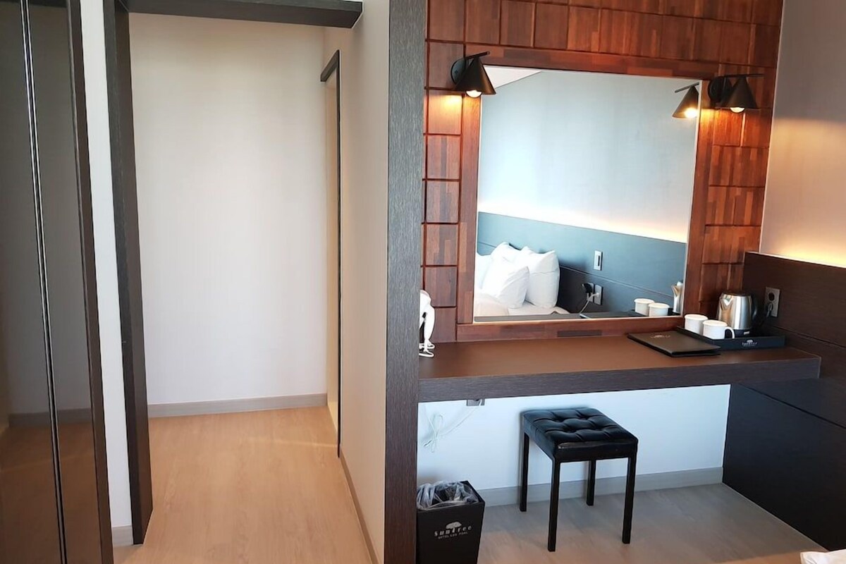 A modern bathroom with a wooden vanity, a mirror, a black trash bin, and a black cushioned stool.