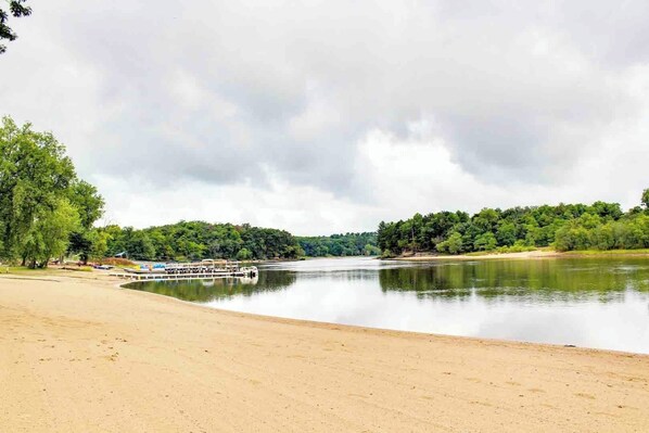 Uma praia arenosa com um cais e barcos, cercada por árvores verdes densas e um céu nublado.