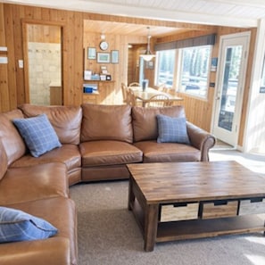 A cozy room with a brown leather sofa, a wooden coffee table, and a wooden wall with framed pictures. - Pine Cottage on Blass Lake (Wisconsin Dells)