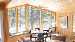A cozy cabin interior with a large wooden dining table, leather sofa, and a window seat overlooking a forest. - Pine Cottage on Blass Lake (Wisconsin Dells)