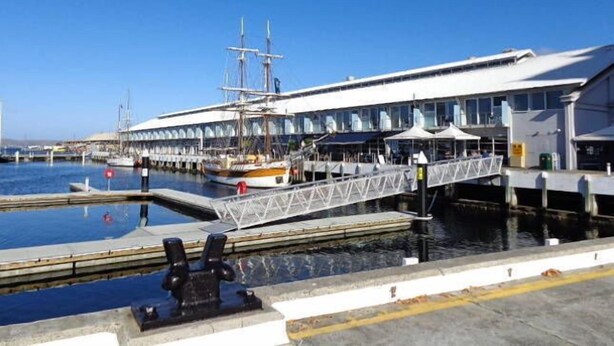 A waterfront building with a large glass facade, a bridge leading to a dock, and a ship moored nearby.