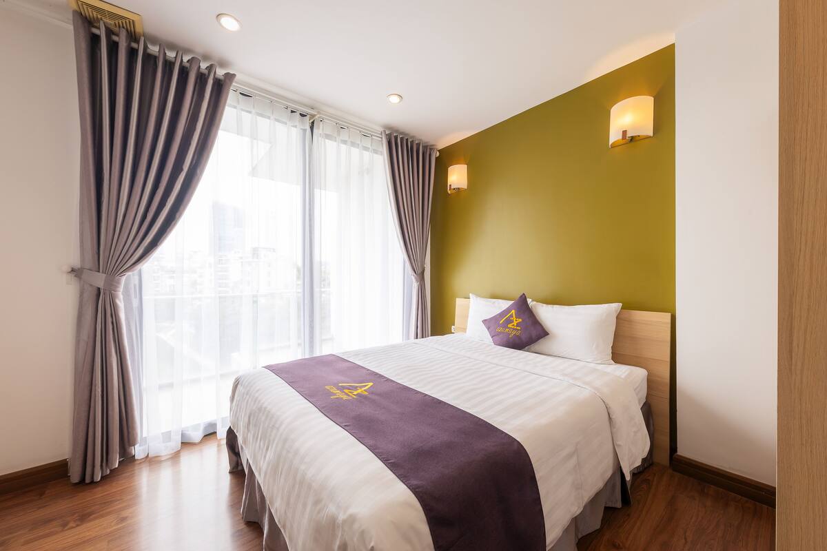 Une chambre d’hôtel avec un grand lit, une tête de lit dotée d’un luminaire et une fenêtre avec des rideaux.