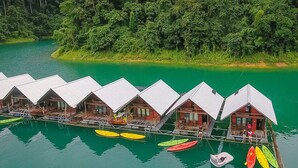 Una hilera de chalets de madera con balcones sobre el agua, unidos por un muelle, rodeados de aguas verdes tranquilas y un bosque denso.