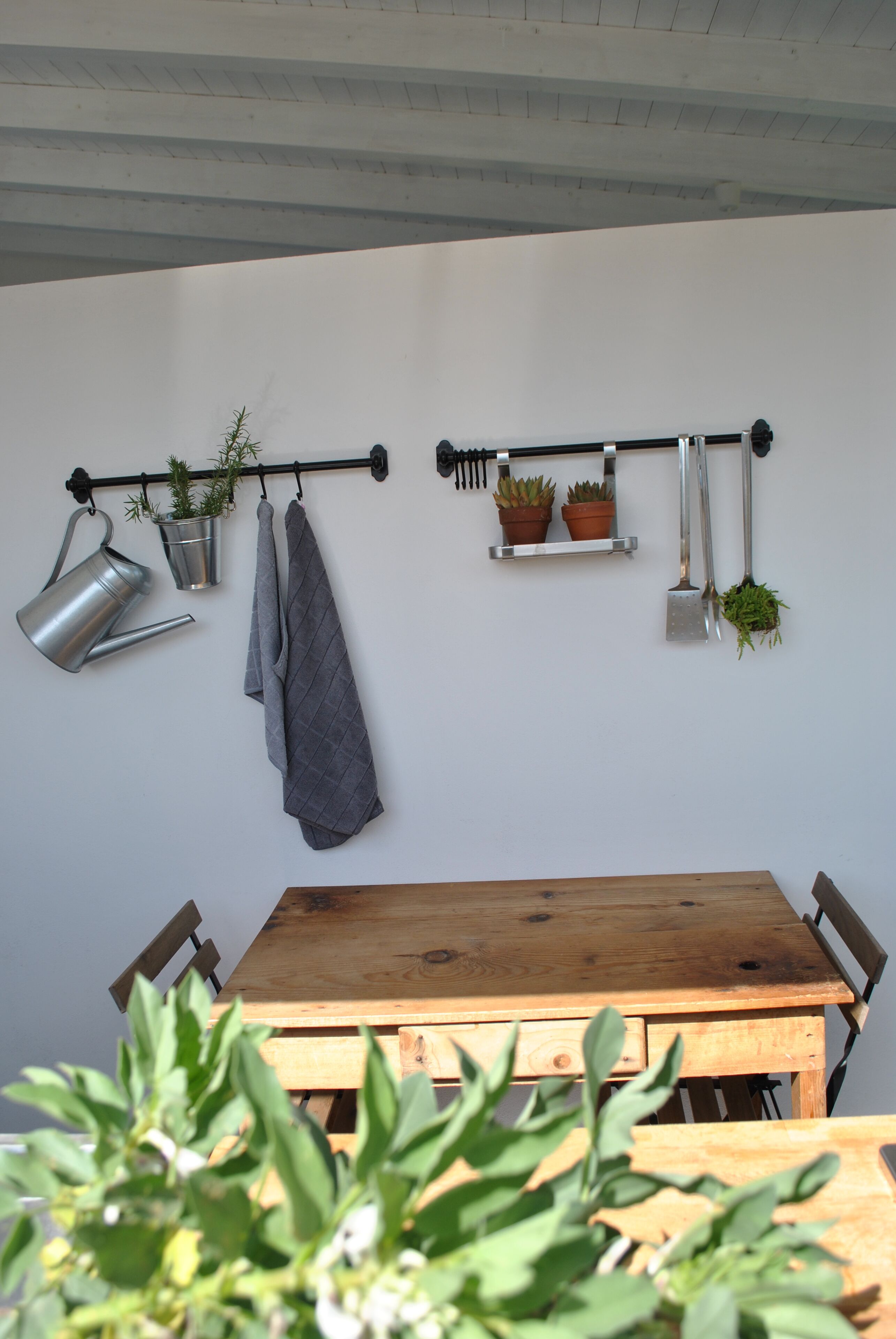 Une table en bois avec des chaises, un arrosoir en métal, une serviette grise et des plantes en pot disposées sur une étagère fixée au mur.