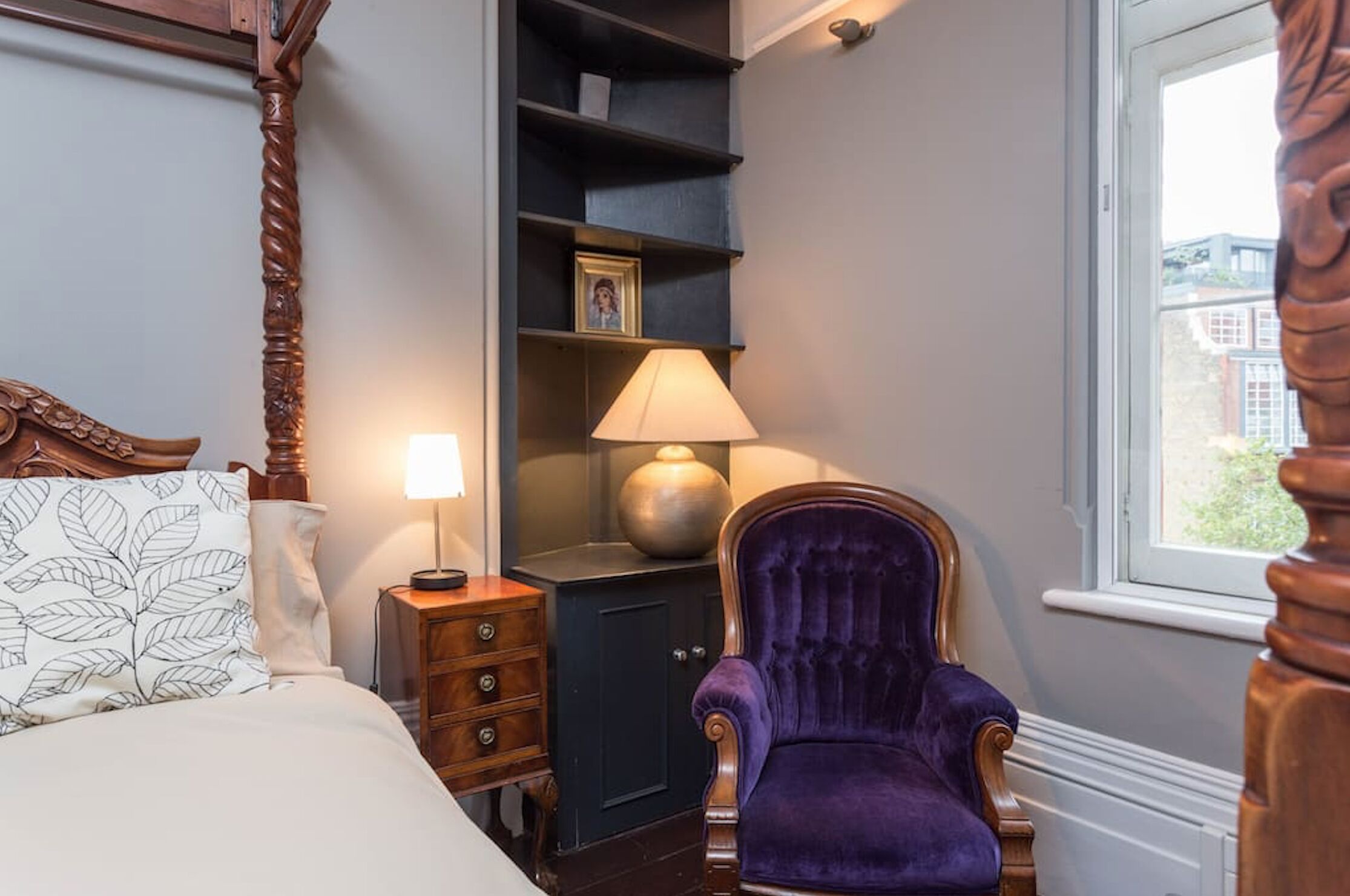 A bedroom with a bed, a wooden nightstand, a dresser, a purple armchair, a bookshelf, and a window with a view of buildings.