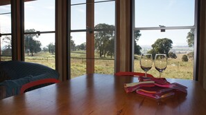 A room with a wooden table, two wine glasses, and a view of a grassy field through large windows. - Idyllic Farm Cottage with Outdoor Bath Under the Stars (Manildra)