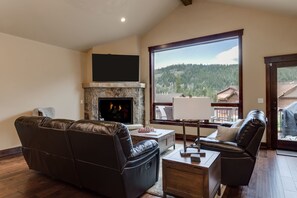 A living room with a fireplace, leather sofas, a coffee table, and a large window offering a view of a forested area. - Luxury Home, Private Hot Tub, Enjoy Keystone, Breckenridge, A Basin, Copper! (Keystone)