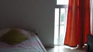 Une chambre à coucher avec un lit, une fenêtre ornée de rideaux rouges, un tableau accroché au mur et un ventilateur de plafond.