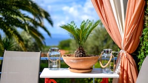 Une table ornée d’un pot de fleurs, de deux bouteilles en verre et d’un pichet en métal, placée sur un balcon d’où l’on peut admirer des palmiers et un ciel dégagé.
