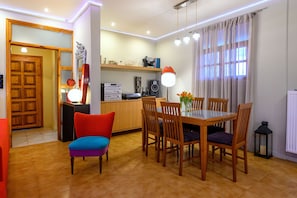 Un coin repas avec une table et des chaises en bois, une chaise rouge, une petite table avec une lampe et une fenêtre avec des rideaux.