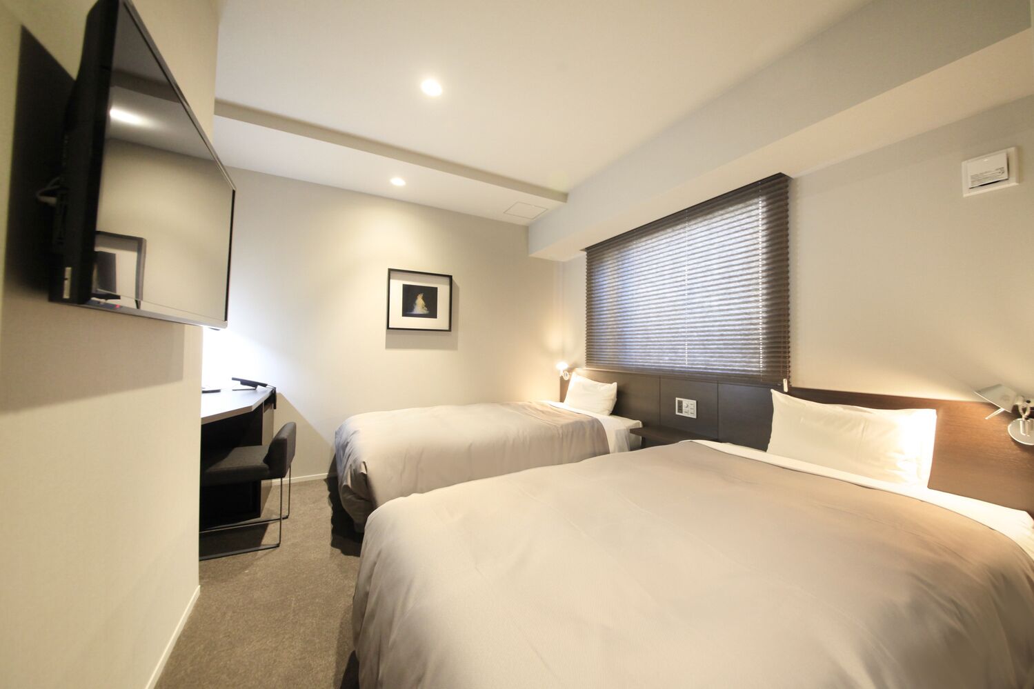 Een hotelkamer met twee bedden, een bureau, een stoel, een televisie, een ingelijst kunstwerk aan de muur en een raam met luxaflex.