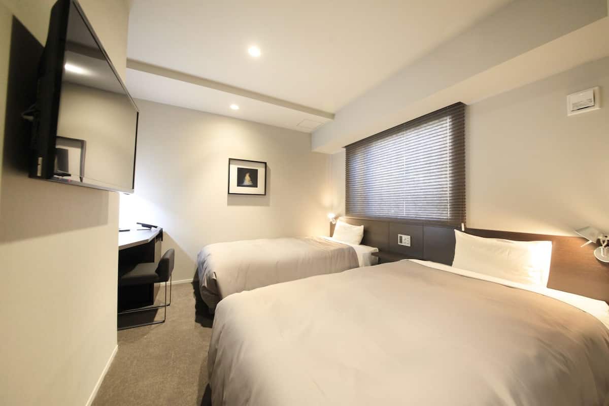 A hotel room with two beds, a desk, a chair, a TV, a framed picture on the wall, and a window with blinds.