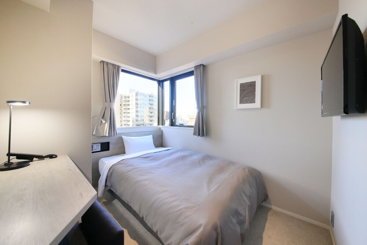Habitación de hotel con cama, escritorio, lámpara, ventana con cortinas y un cuadro en la pared.