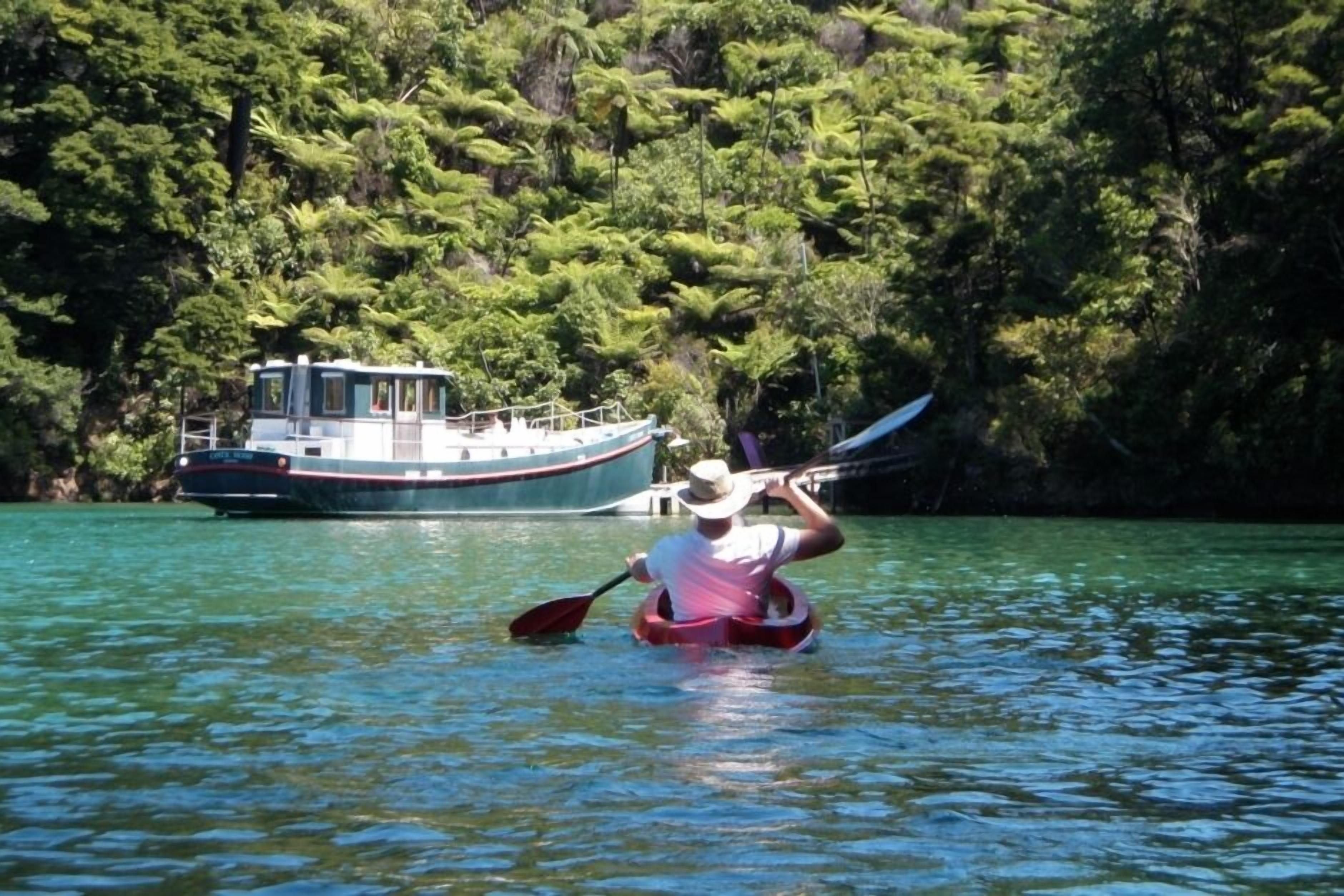 Una persona en kayak en un río tranquilo, con una embarcación al fondo y abundante vegetación en las orillas.