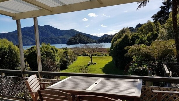 A covered outdoor seating area with a table and chairs, overlooking a lake and mountains. - Relax and Enjoy - Waterfront Bach in Ngakuta Bay (Ngakuta Bay)