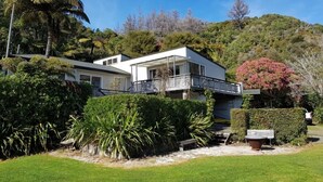 A modern two-story house with a balcony, surrounded by lush greenery and a hillside. - Relax and Enjoy - Waterfront Bach in Ngakuta Bay (Ngakuta Bay)