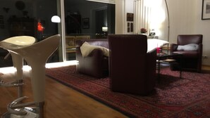 A modern living room with a large window, a dark brown sofa, a white chair, and a red patterned rug.
