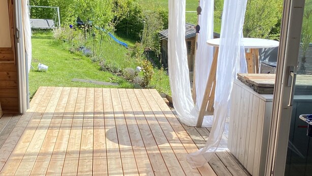 A room with a wooden deck, a view of a lush garden, and a large window with white curtains.