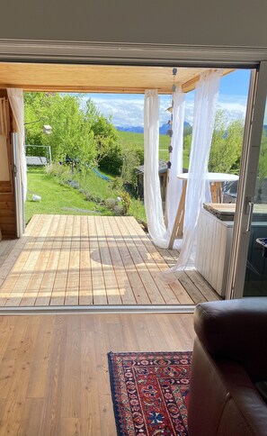 Ein Zimmer mit Holzbalkon, Blick auf einen üppigen Garten und einem großen Fenster mit weißen Vorhängen.