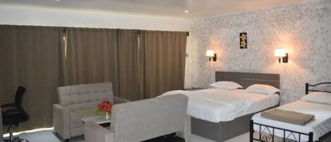 Habitación de hotel con dos camas, un sofá, un escritorio y una silla. Cuenta con lámparas de pared y una ventana con cortinas.