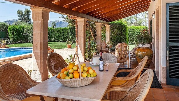 Un área de comedor al aire libre techada con sillas de mimbre, una mesa con una cesta de frutas, una botella y vasos.