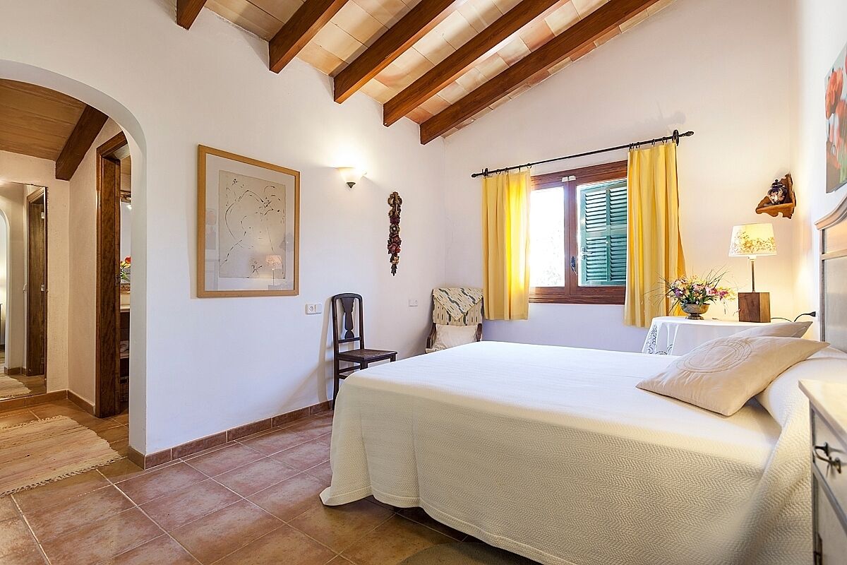 A bedroom with a large bed, a window with yellow curtains, a wall-mounted lamp, and a framed picture.