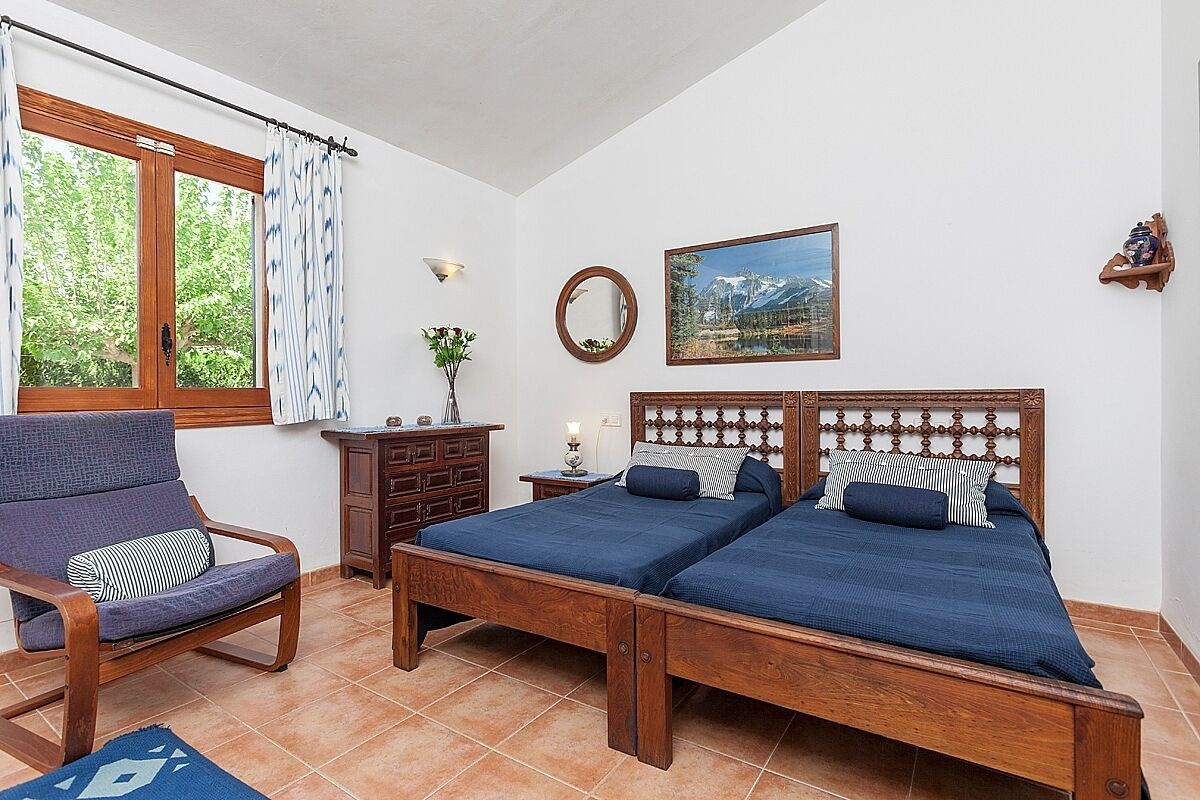 Una camera da letto con un letto di legno, una sedia, una cassettiera, una finestra con tende e uno specchio fissato al muro, oltre a un quadro incorniciato.