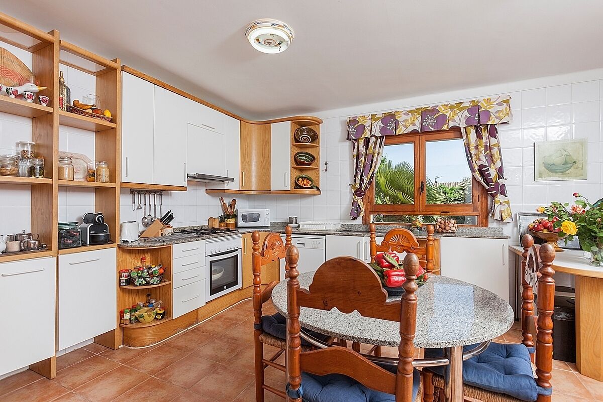 A kitchen with white cabinets, wooden shelves, a round table with chairs, and a window with curtains.