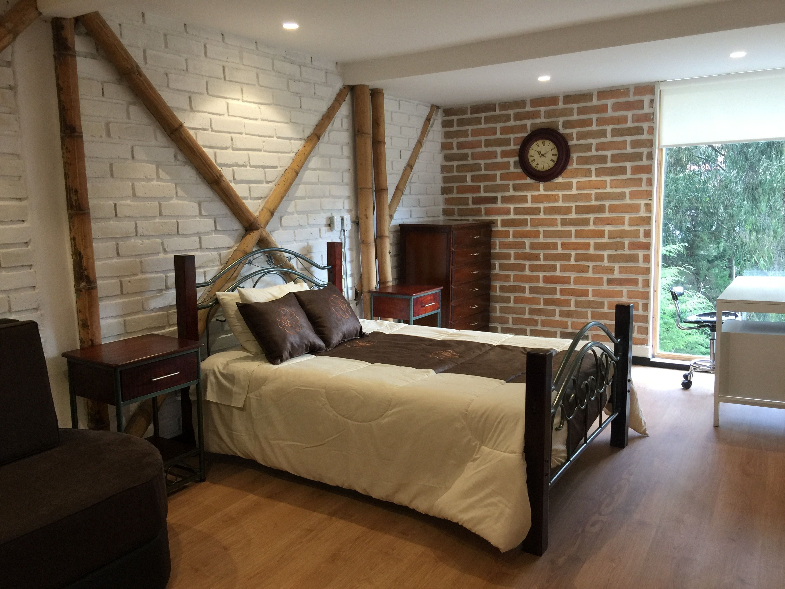 A bedroom with a bed, wooden beams, a brick wall, a clock, and a window with a view of greenery.