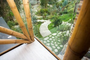 A view through a window with bamboo frames, a stone pathway, and a lush garden with various plants and a small pond. - Bambu Sierra (Cuenca)