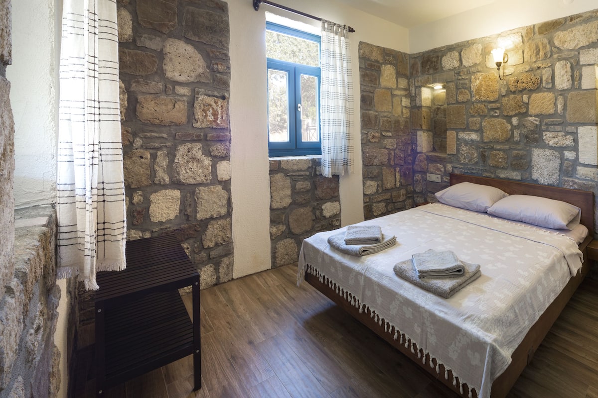A bedroom with a stone wall, a bed with towels, a window with curtains, and a nightstand.
