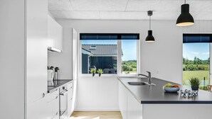 A modern kitchen with a large island, a window with a view of a house and greenery, and a black pendant light. - Lovely home in Glesborg with sauna (Glesborg)
