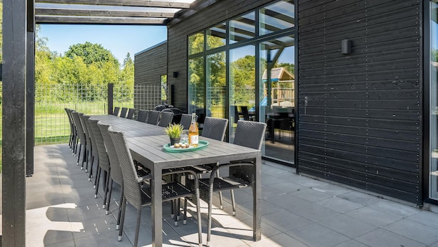 An outdoor patio with a long table and chairs, a grill, and a glass enclosure with a view of greenery. - Lovely home in Glesborg with sauna (Glesborg)