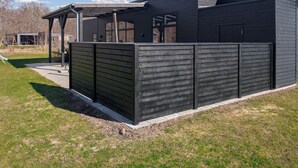 A modern building with a dark exterior, a covered patio area, and a fenced outdoor space. - Lovely home in Glesborg with sauna (Glesborg)
