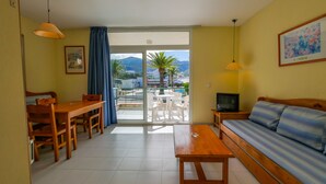A living room with a sofa, a wooden table, a TV, and a balcony with a view of palm trees and buildings. - Rescator Resort 322 - One Bedroom Apartment, Sleeps 4 (Roses)