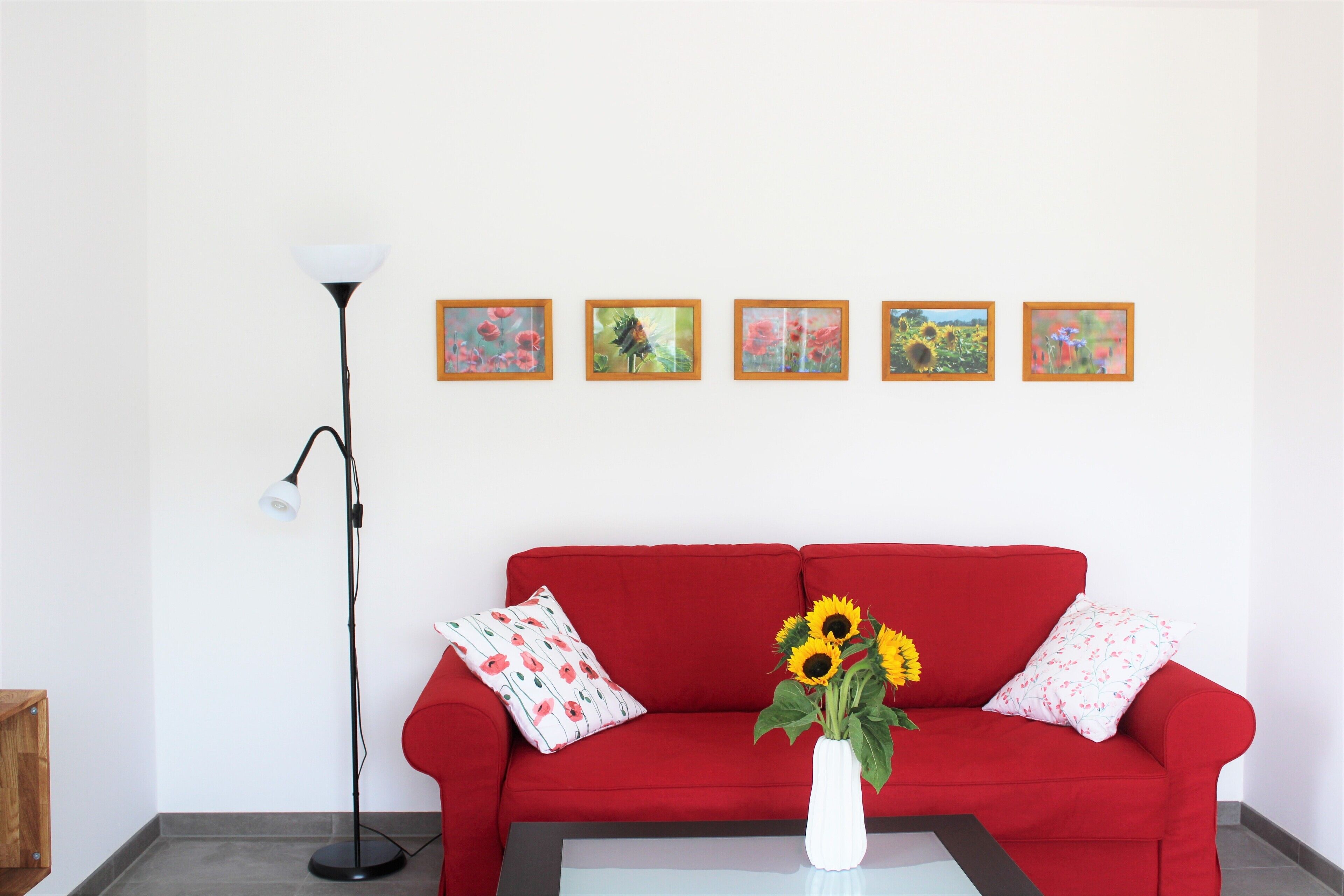 Un sofá rojo con un jarrón blanco lleno de girasoles, una lámpara de pie y cinco cuadros enmarcados en la pared.