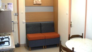 Una habitación compacta con kitchenette, un sofá con cojines, una mesita y una puerta.
