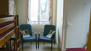 Una habitación con una cama superpuesta, dos sillones con cojines, una ventana con cortinas y una puerta.