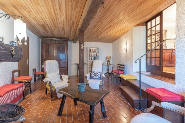 A cozy room with wooden beams, a fireplace, a bookshelf, and a dining area with chairs and a table.