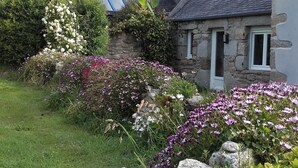 Una casa de piedra con techo inclinado y un pequeño jardín con diversas flores y una gran roca.