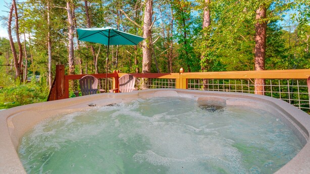 Un jacuzzi rodeado por una terraza de madera, con una silla de madera y una sombrilla verde cerca.