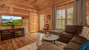 Interior de una cabaña de madera con un televisor grande, un sofá, una mesita de centro redonda y una silla.
