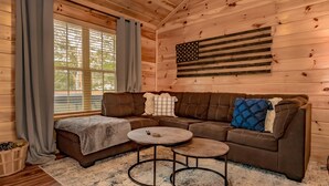 Un interior de cabaña acogedor con detalles de madera, un sofá modular, una pequeña mesa redonda y un ventanal con cortinas.
