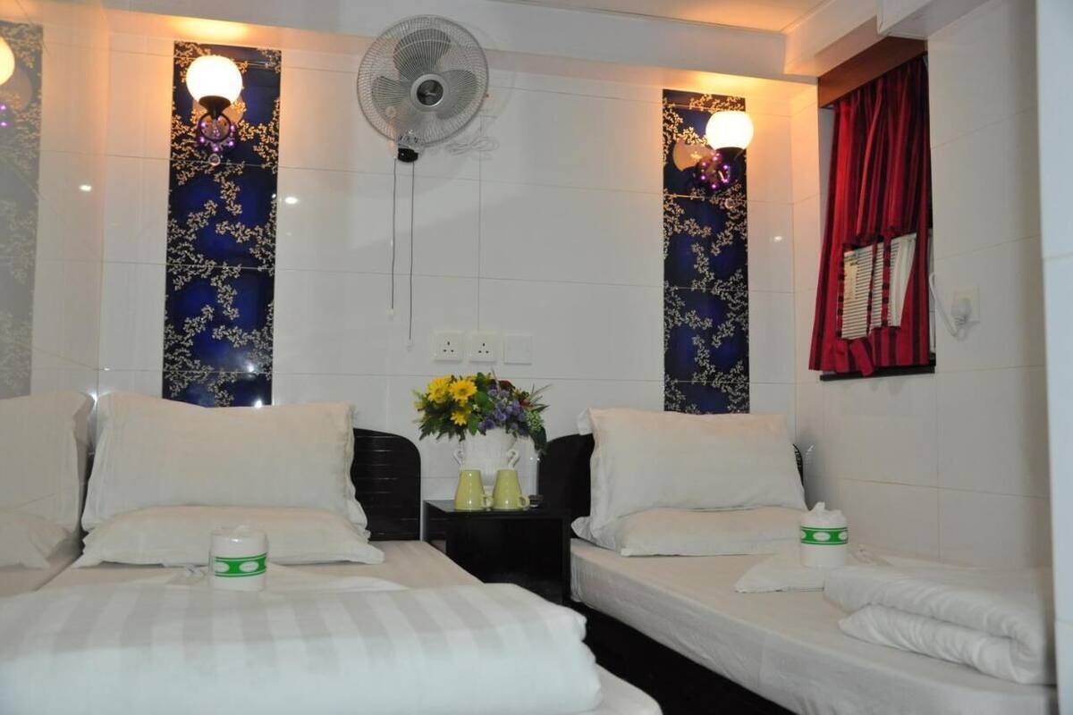 A hotel room with two beds, a standing fan, decorative wall panels, and a window with red curtains.